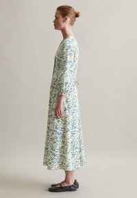 Floral midi Kleid in Weiß mit grünen und blauen Mustern, Glockenärmeln und einer taillierten Taille. Kombiniert mit schwarzen Ballerinas.