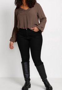 Blouse marron à manches longues avec un col en V et une texture plissée, associée à un jean noir taille haute et des bottes en cuir noires jusqu'aux genoux.