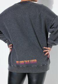 Sudadera grande de color gris oscuro con puños acanalados, que presenta un estampado desvanecido "PODREMOS HABLAR DESPUÉS" en un degradado de naranja a morado en el dobladillo.