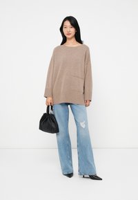 Przesadny beżowy sweter z kieszenią z przodu, zestawiony z jasnoniebieskimi dżinsami w stylu distressed oraz czarną torebką na sznurek. Czarne buty na wysokim obcasie.