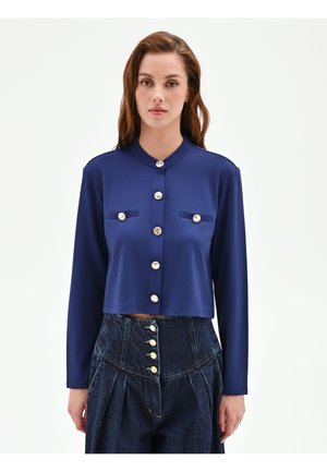 Donna che indossa un cardigan corto blu navy con bottoni e due tasche sul petto e pantaloni in denim scuro a vita alta con molteplici bottoni.