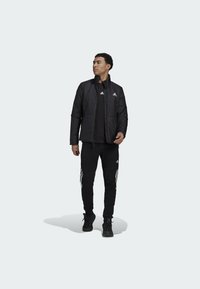 Giacca imbottita nera con colletto alto e logo ricamato, abbinata a una camicia nera e pantaloni sportivi neri con strisce bianche.