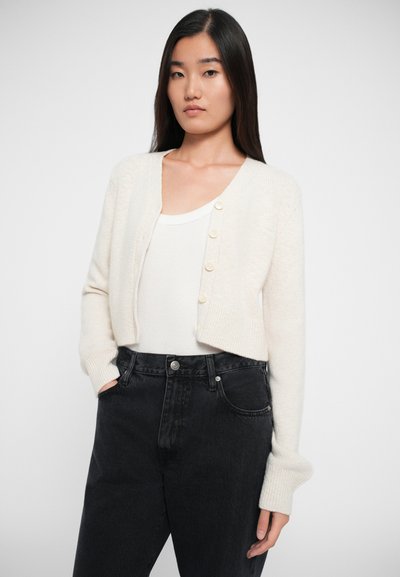 Kremfarget, kort cardigan laget av mykt materiale, med lange ermer, rund hals. Den har fem knapper foran.