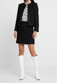 Svart tweedjacka med texturerad yta, kombinerad med en vit blus med stjärnmönster och en matchande svart kjol, kompletterad med glansiga vita knähöga stövlar.