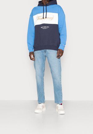 Sudadera con capucha de bloques de color azul y azul marino con un logo gráfico, combinada con vaqueros de lavado claro y zapatillas blancas, con una tela texturizada.