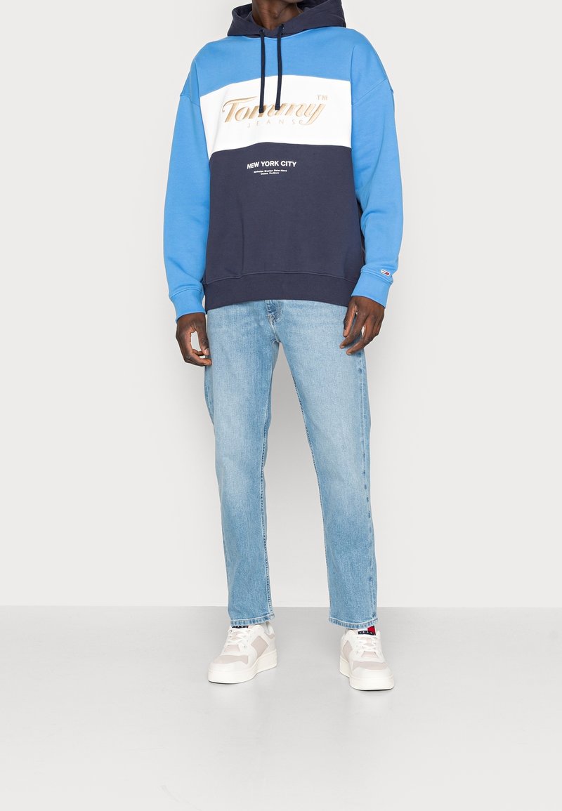 Modrá a tmavě modrá mikina s grafickým logem, doplněná světle vyšisovanými džíny a bílými teniskami, vyrobená z texturované látky.