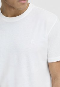 Bílý krátký rukáv tričko s kulatým výstřihem a jemným vyšívaným logem na hrudi. Materiál je lehký a hladký.