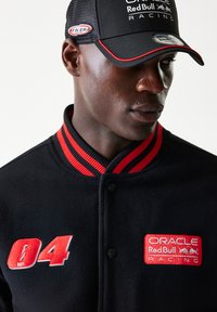 Μαύρο μάλλινο σακάκι με κόκκινο ριγέ γιακά, διακοσμημένο με κόκκινη κεντητική, αυτοκόλλητα με λογότυπο και μαύρα κουμπιά τύπου σναπ. Μαύρο καπέλο με λογότυπο και διακοσμητικά στοιχεία.