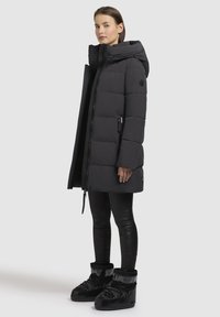 Cappotto imbottito nero con design trapuntato, cappuccio grande e tasche con zip, abbinato a leggins neri e stivali invernali robusti.