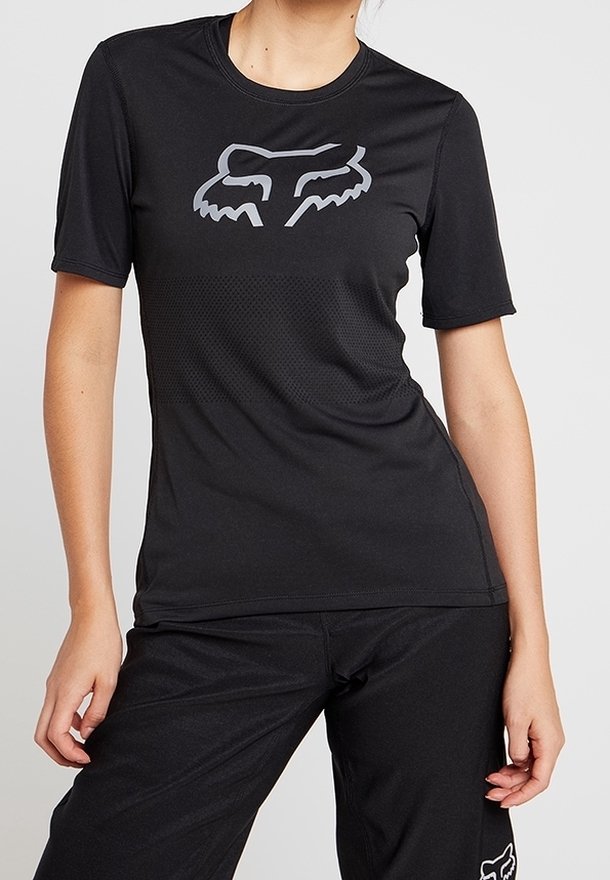 Frau trägt ein enganliegendes schwarzes kurzärmeliges Sportshirt mit einem grauen stilisierten Fuchskopf-Logo auf der Brust, kombiniert mit schwarzen Hosen.