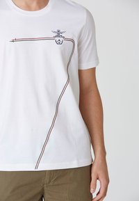 T-shirt bianco in cotone con bordo diagonale rosso, verde e nero, con ricamo di aquila e dettaglio del logo sul petto.