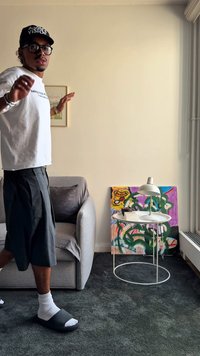 Ένα άτομο με λευκό μπλουζάκι και σκούρο σορτς στέκεται κοντά σε ένα γκρι καναπέ, μια λευκή επιδαπέδια λάμπα και έναν πολύχρωμο αφηρημένο πίνακα στο πάτωμα.