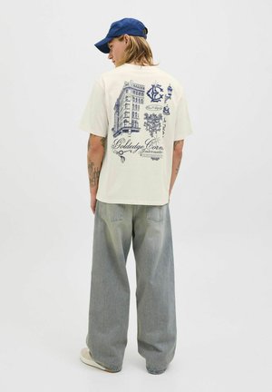 Flødefarvet bomulds-t-shirt med blå grafisk tryk af en bygning og dekorative elementer på ryggen. Matchet med løse grå denimbukser.