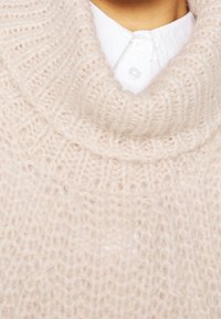 Helles rosa Strickpullover mit oversized Kragen, weichem Material und Rippmuster. Über einem weißen Hemd mit Knopf getragen.