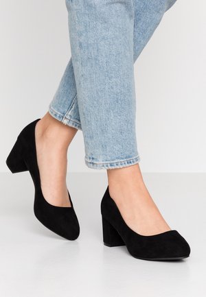 Person iført lyseblå jeans og sorte ruskind blokhælssko står på en hvid gulv mod en ensfarvet baggrund.