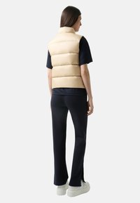 Beige gepolsterte Weste mit Stehkragen und horizontalen Nähten. Kombiniert mit schwarzen Hosen und weißen Sneakers für ein komplettes Outfit.