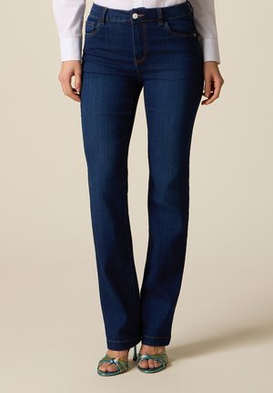 Donna che indossa jeans blu scuro a gamba dritta e sandali con tacco alto multicolori con punta aperta, in piedi su uno sfondo neutro.