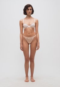 Kobieta stojąca boso, ubrana w beżowy koronkowy biustonosz i dopasowaną bieliznę, na jednolitym jasnym tle.