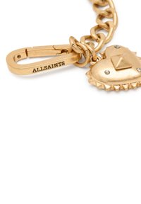 Charm de metal color dorado adjunto a un eslabón de cadena, con forma de corazón, un tachuela en forma de pirámide y dos pequeñas piedras transparentes. Presenta un grabado de marca.