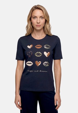 Femme portant un t-shirt noir avec des imprimés de cœurs et de lèvres de différentes couleurs et le texte "hugs and kisses" sur le devant.