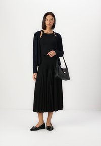 Jupe midi plissée noire associée à un haut noir ajusté et un cardigan bleu marine. Sac à main noir avec des accents dorés, et des chaussures plates noires avec des embellissements.