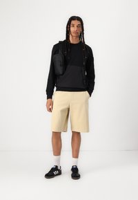 Svart hettegenser med halv glidelås og lommeinnlegg, sammen med beige shorts. Modellen har på seg svarte sneakers med hvite striper og grå sokker.