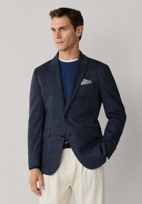 Muž v tmavě modrém saku s jemným károvaným vzorem, modrém svetru, bílých kalhotách a vzorovaném kapesníku, stojící s jednou rukou v kapse.