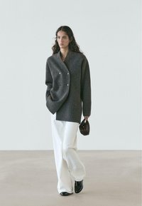 Cappotto doppiopetto grigio in lana con vestibilità oversize, indossato sopra una maglia crema e pantaloni bianchi larghi. Modella che tiene una piccola borsa marrone.