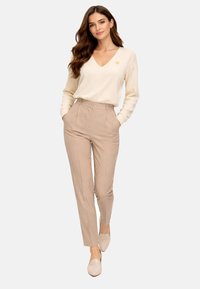 Femme aux cheveux longs et ondulés portant un pull crème à col en V et un pantalon beige taille haute, debout les mains dans les poches sur fond blanc.