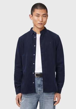 Junger Mann, der ein marineblaues Hemd mit Knöpfen über einem weißen T-Shirt trägt, mit hellblauen Jeans und schwarzem Gürtel, vor einem einfarbigen Hintergrund stehend.