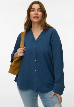 Chemise ample à manches longues de couleur sarcelle avec devant boutonné, tissu texturé et boutons dorés. Associée à un jean bleu clair déchiré et un sac beige.