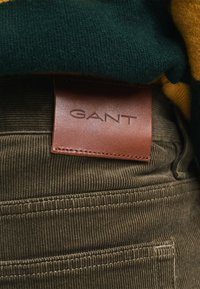 Brązowe spodnie z korduroju mają skórzaną metkę z wytłoczonym logo "GANT". Powierzchownie widoczny jest zielono-żółty sweter w paski.