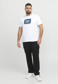 Valge puuvillane t-särk sinise graafilise trükiga, kus ees on kirjas "JACK & JONES", koos mustade cargo-pükstega ja valgete tossudega.