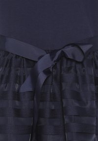 Abito blu scuro con vita a strisce orizzontali testurizzate e un nastro blu lucido annodato in un semplice fiocco.