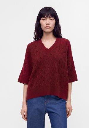 Kvinde med langt sort hår iført en rød kabelstrikket kortærmet sweater og blå jeans, stående mod en ensfarvet hvid baggrund.