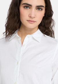 Camisa blanca de botones con un cuello clásico, tapeta delantera con botones y un sutil logo bordado en el lado izquierdo del pecho. Textura de tela suave.