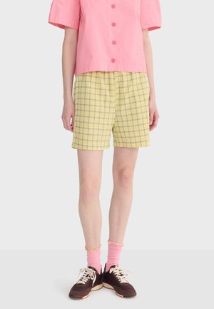 Osoba oblečená v ružovej košeli s krátkym rukávom na gombíky, žlté kárované šortky, ružové ponožky a tmavé tenisky stojaca pred jednofarebným pozadím.