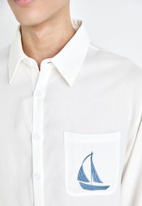 Hvit skjorte med knapper, med spiss krage og lomme på venstre bryst med blå seilbåtbroderi. Mykt, lett stoff.