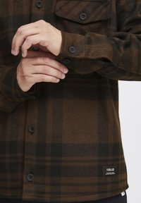 Chemise écossaise marron en tissu doux, avec des poignets à boutons et une poche poitrine avec des boutons noirs. L'étiquette sur l'ourlet indique "ISOLID."