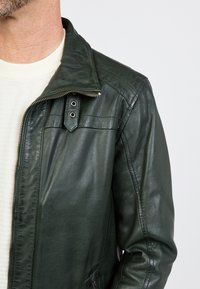 Man in een donkergroene leren jas over een witte, gestructureerde trui, weergegeven van de nek tot het midden van de romp op een lichte achtergrond.