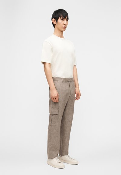 Άνδρας που στέκεται, φοράει κρεμ κοντομάνικη μπλούζα, μπεζ παντελόνι cargo με πλαϊνές τσέπες και κρεμ αθλητικά παπούτσια, σε απλό λευκό φόντο.