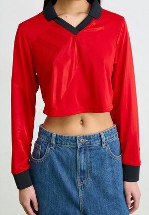 Rotes langärmeliges, kurz geschnittenes Shirt mit schwarzem Kragen und Bündchen, kombiniert mit hoch taillierter blauer Denim-Jeans, der freiliegende Bauch ist sichtbar.