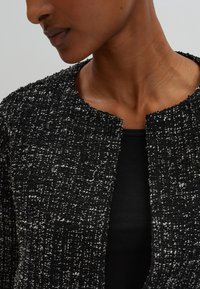 Veste en tweed texturée noir et blanc avec un col rond, devant ouvert et une silhouette cintrée, associée à un haut noir en dessous.