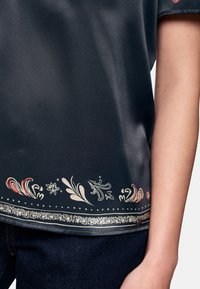Dark satin blouse featuring a floral design in red and cream along the hem, with a metallic accent on the border and short sleeves.