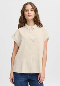 Camicetta beige in lino a maniche corte con patta bottonata e colletto a punta, caratterizzata da una vestibilità ampia e una superficie leggermente testurizzata.