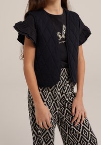 Czarna pikowana kamizelka z okrągłym dekoltem noszona na czarnym t-shircie z złotymi detalami i wzorem zwierzęcym. Połączona z wzorzystymi spodniami w czarno-białe pasy.