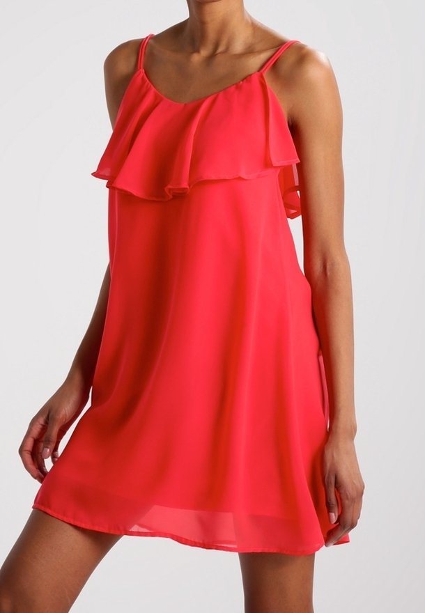 Femme portant une robe rouge vif sans manches avec un col à volants et de fines bretelles, debout devant un fond uni.