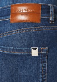 Vaqueros de mezclilla azul oscuro con una etiqueta de cuero marrón en la cinturilla que dice "WEEKEND MaxMara." Incluye una pequeña etiqueta blanca con un diseño de mariposa negra.