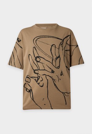 Mężczyzna ubrany w beżowy oversize t-shirt z czarnymi ręcznymi szkicami, czarne szerokie spodenki do kolan i czarne botki na kostkę, stojący na jednolitym tle.