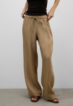 Persoon draagt losse zandkleurige linnen broek met trekkoord in de taille en bruine sandalen met bandjes, staand tegen een effen grijze muur.